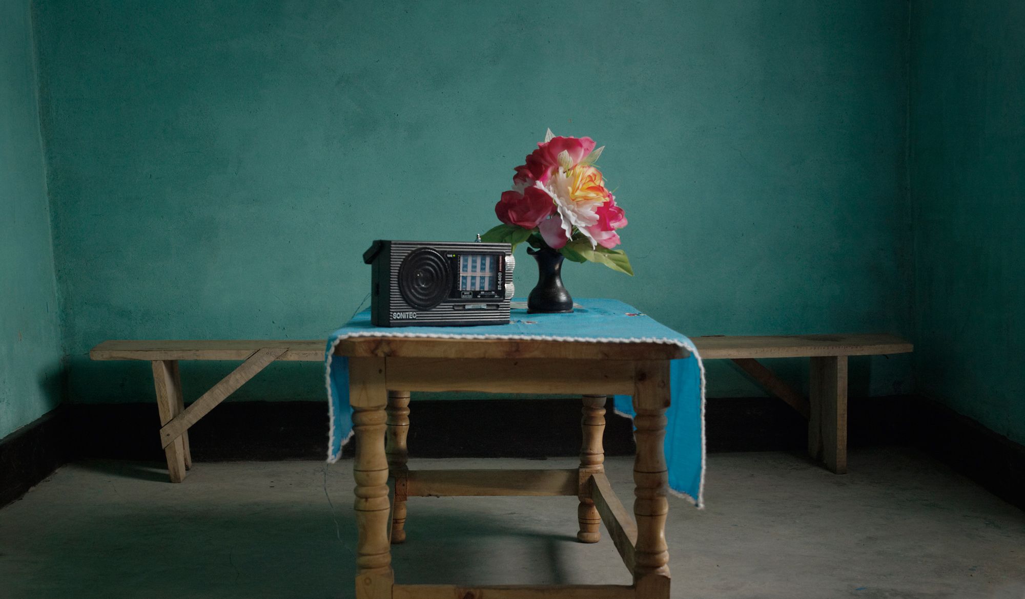 A portable radio and a vase of flowers on a wooden table against a turquoise wall in Rwanda
