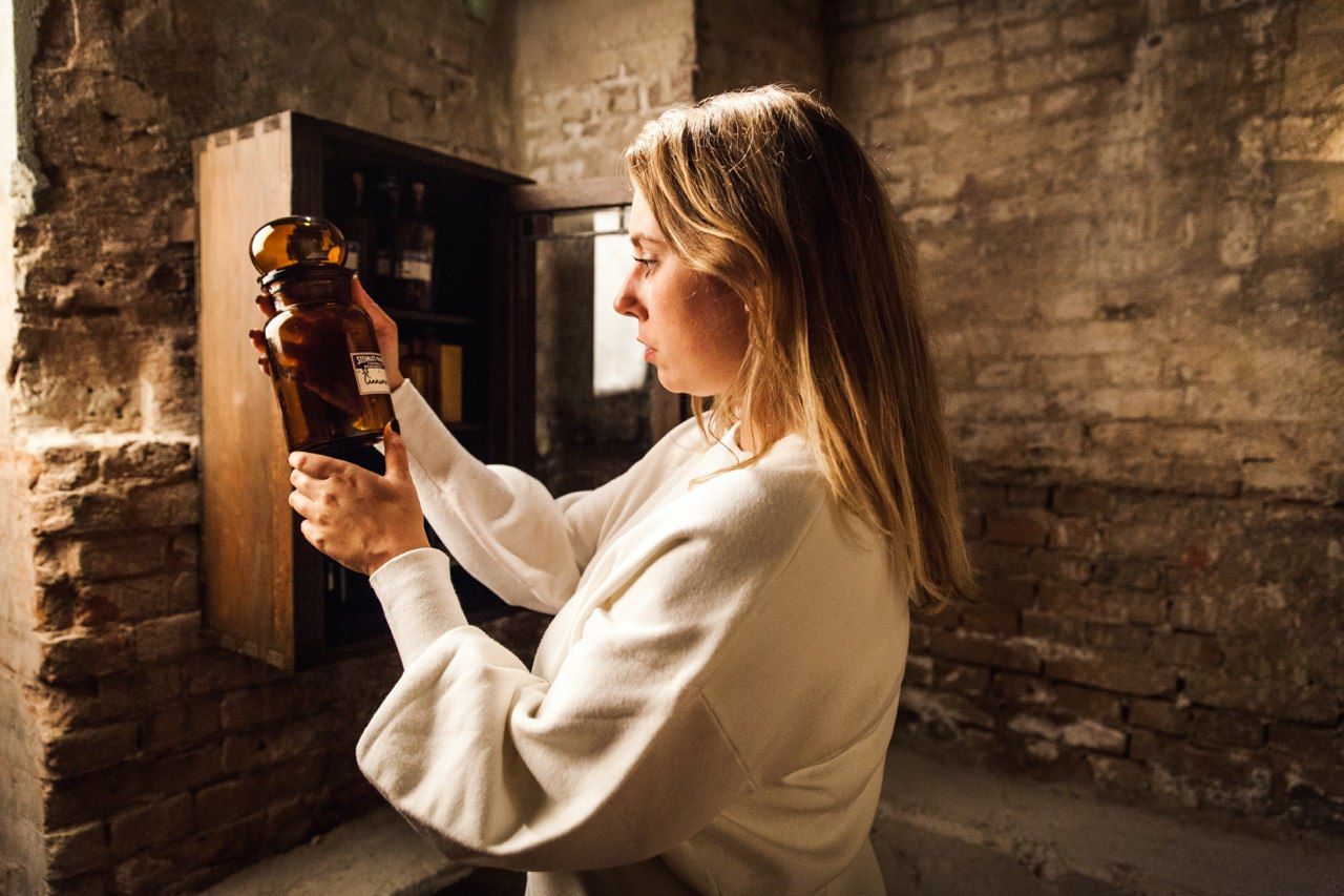 A woman in a dark brick room studies an amber bottle taken from a wooden cabinet during an escape room